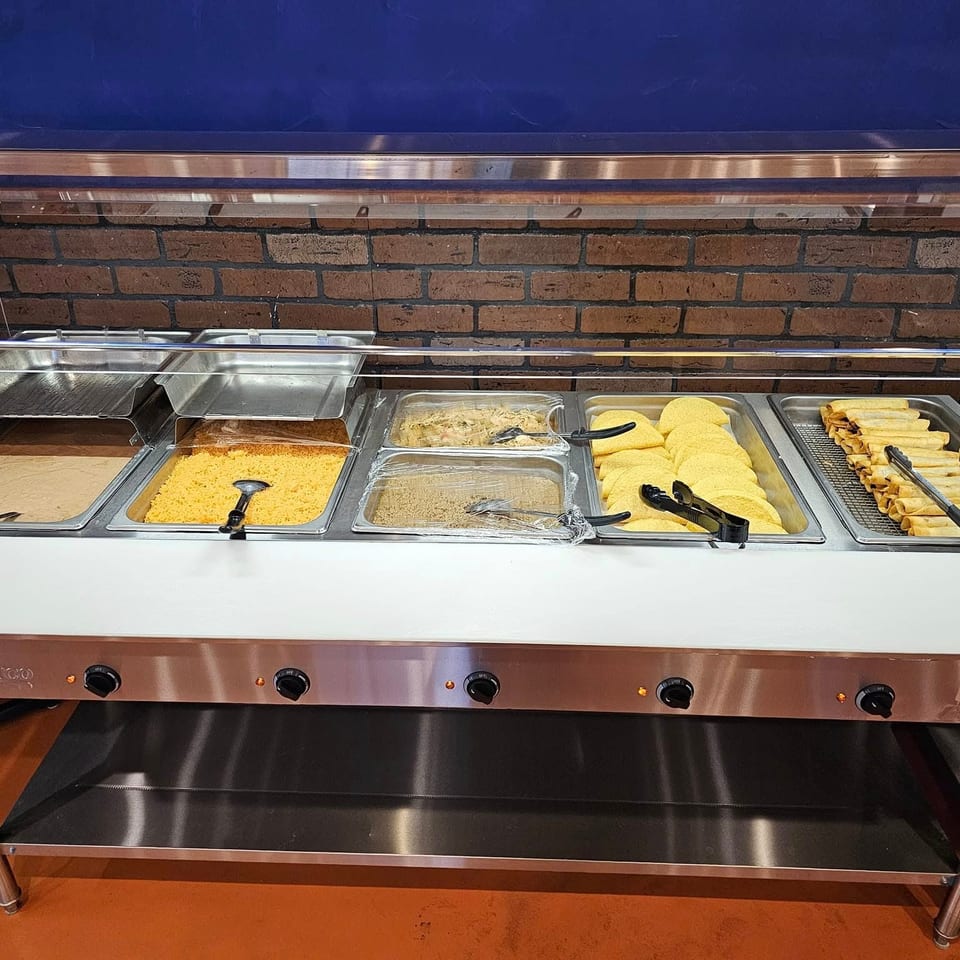 A buffet line with various Mexican dishes, including rice, tortillas, and taquitos, against a brick wall.