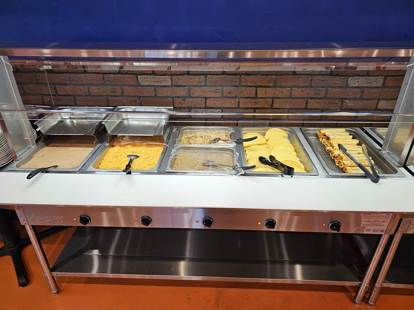 A buffet line with various Mexican dishes, including rice, tortillas, and taquitos, against a brick wall.